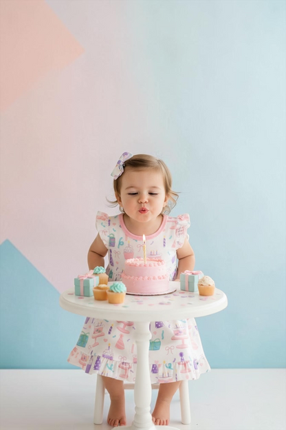 Toddler girl wearing a Nannette's Online Designs birthday dress with ruffle hem and cake print, blowing out a candle.